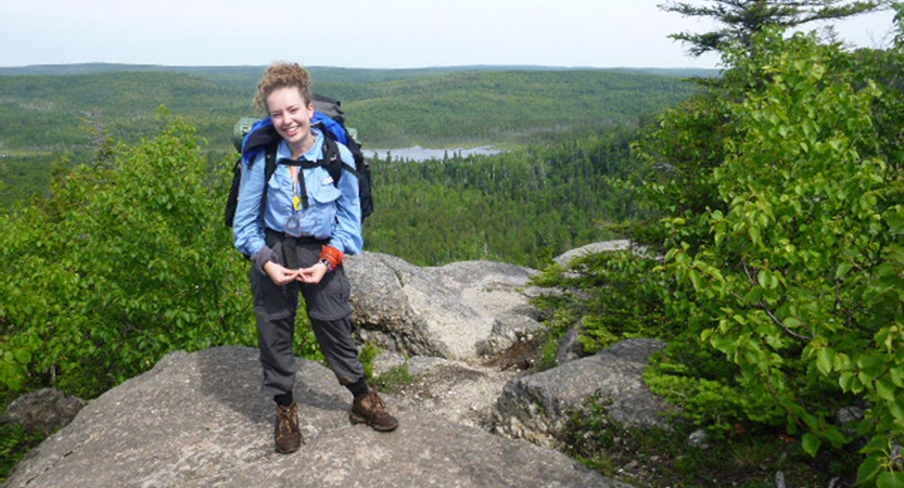 A person wearing a backpack stands on a rocky outlook, smiling at the camera. Behind them stretches a vast green forrest and a body of water. 